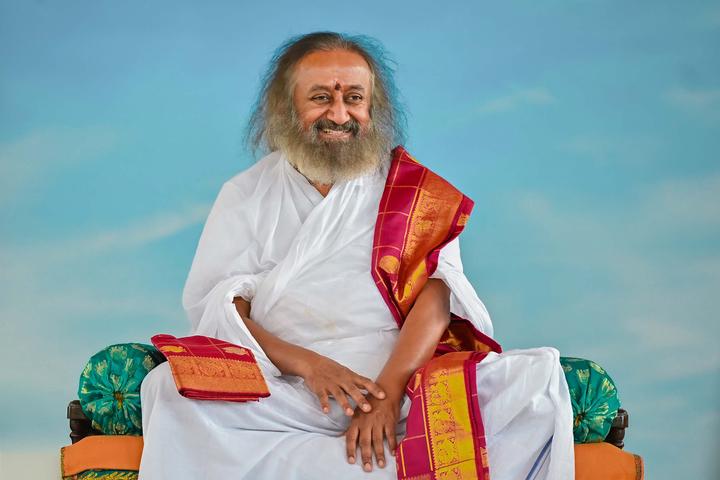 An elderly man with a long gray beard wearing white robes and a red-gold sash sits cross-legged against a clear blue sky, smiling warmly.